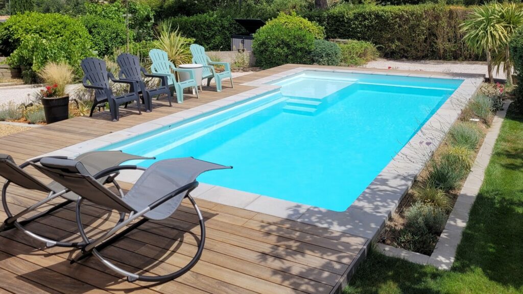 Piscine coque enterrée avec margelles en pierre et terrasse en bois. Aménagements paysagers extérieurs.